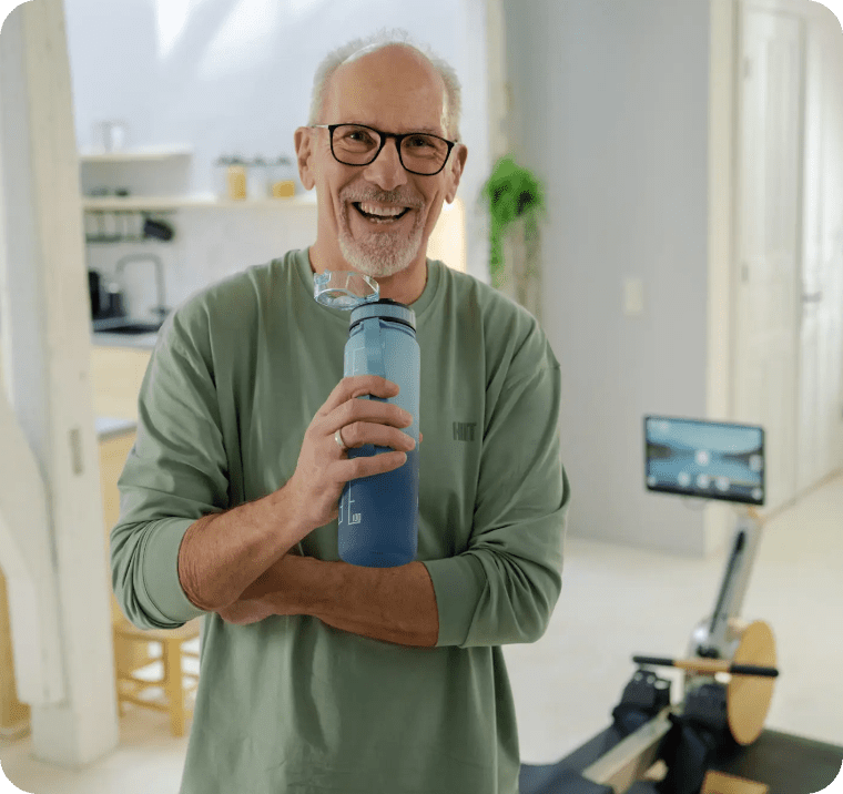 Pulstraining auf dem Rudergerät: So findest du deinen perfekten Trainingspuls 3 „Älterer Mann mit Wasserflasche lächelt vor einem Rudergerät im Wohnzimmer.“