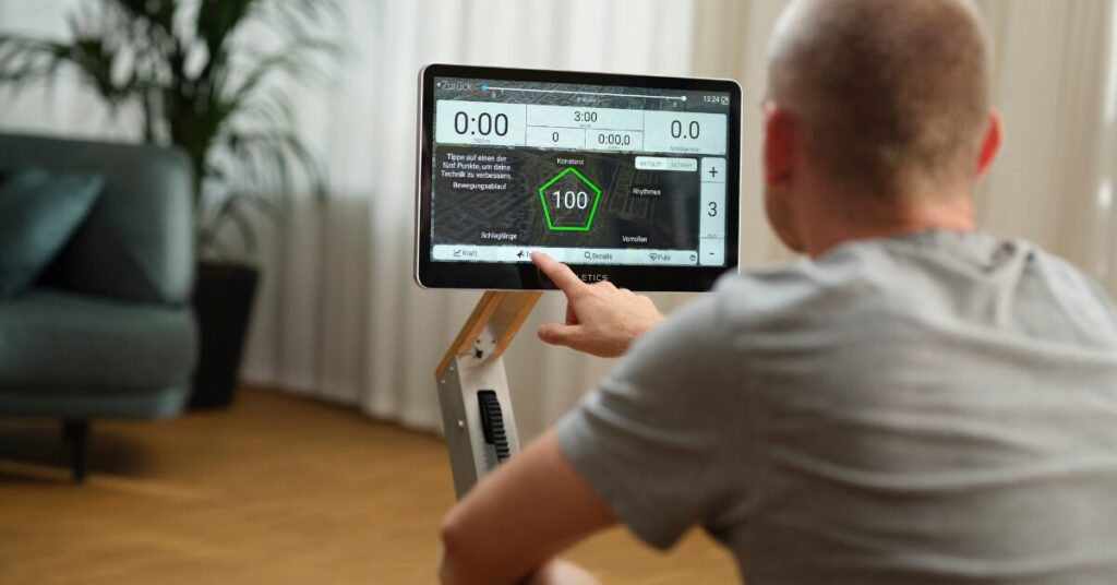 Rudern bei Ischiasschmerzen – Bewegung statt Schonung 4 Mann bedient den Virtuellen Trainer am Display eines Rudergeräts zur Trainingssteuerung in einem Wohnzimmer