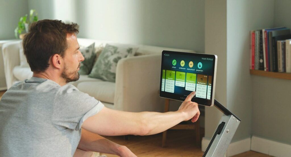 Mann bedient das Touchscreen-Display eines AUGLETICS Rudergeräts, auf dem verschiedene Trainingsprogramme angezeigt werden