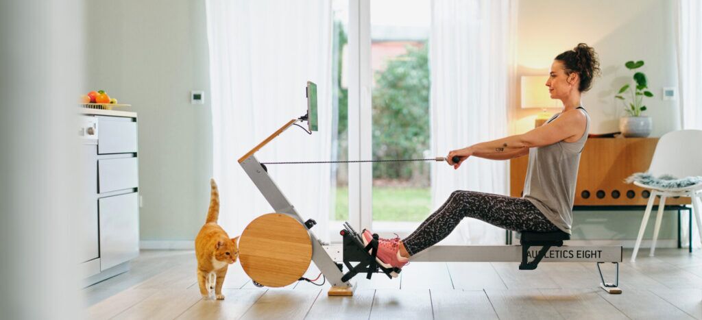 Une femme s'entraîne sur le rameur AUGLETICS, tandis qu'un chat orange se promène à côté.