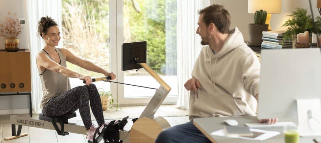 Woman trains on AUGLETICS rowing machine while a man smiles at her from his desk