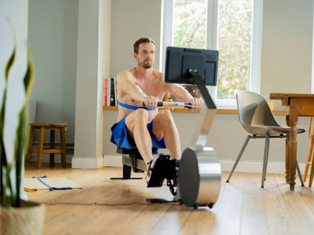 Un homme s’entraîne sur un rameur moderne dans un salon lumineux avec parquet en bois. Il connaît sa fréquence cardiaque maximale et s’entraîne dans la zone anaérobie.