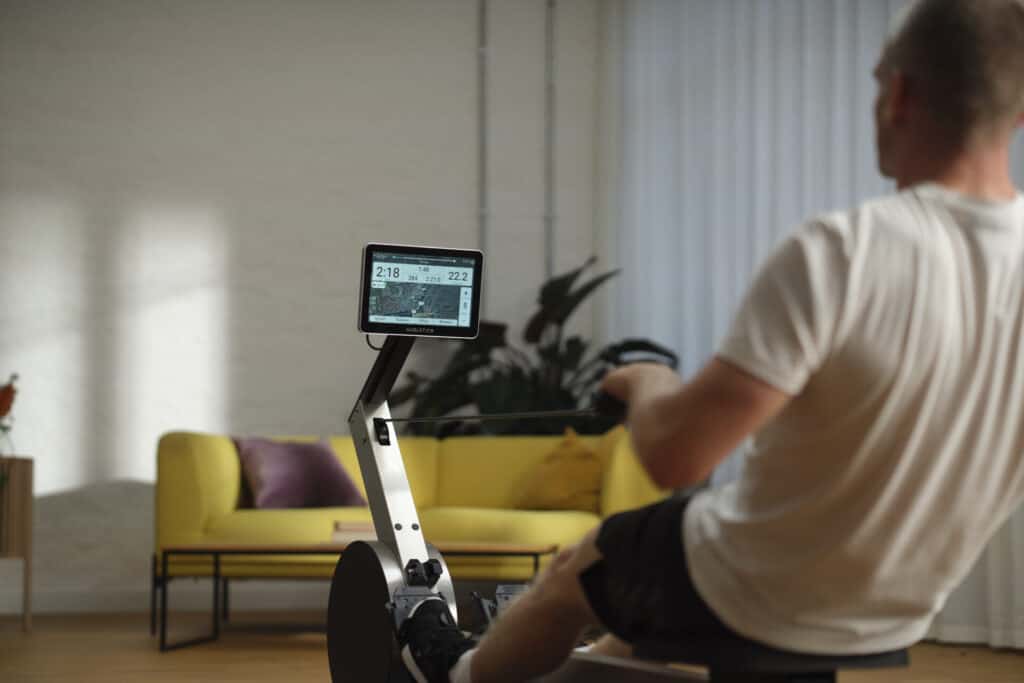 Un homme s'entraîne sur un rameur dans un salon aménagé avec goût, avec des plantes et un mobilier confortable. Une véritable alternative à la salle de sport.