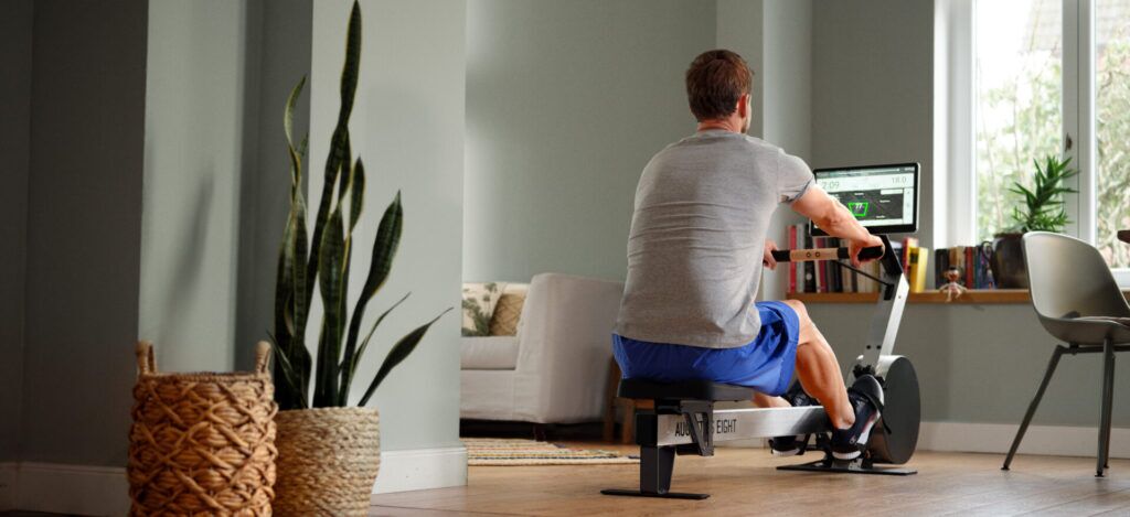 Mann trainiert auf einem Rudergerät in einem stilvoll eingerichteten Wohnzimmer mit Pflanzen und gemütlicher Einrichtung. Eine echte Alternative zum Fitnessstudio.