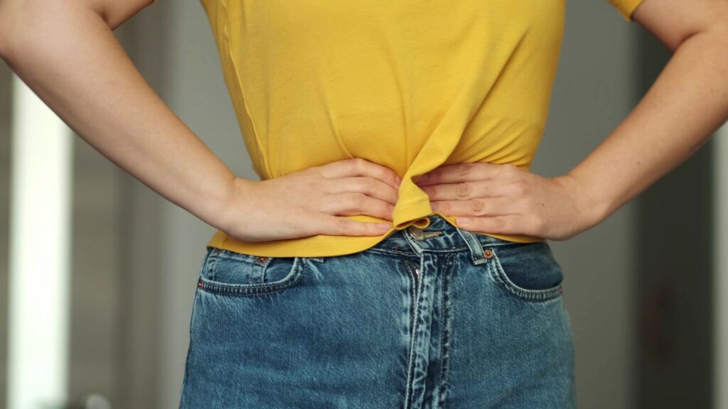 Nahaufnahme eines Menschen, der ein gelbes T-Shirt und blaue Jeans trägt und beide Hände in den unteren Rücken legt
