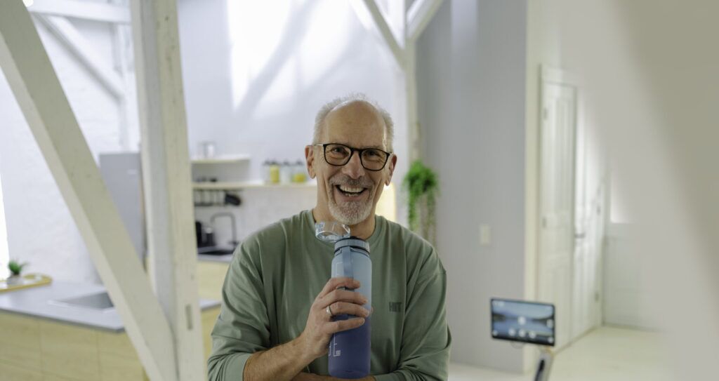 Älterer Mann mit Brille lächelt in einem modernen Raum und hält eine Trinkflasche, im Hintergrund ein Rudergerät.