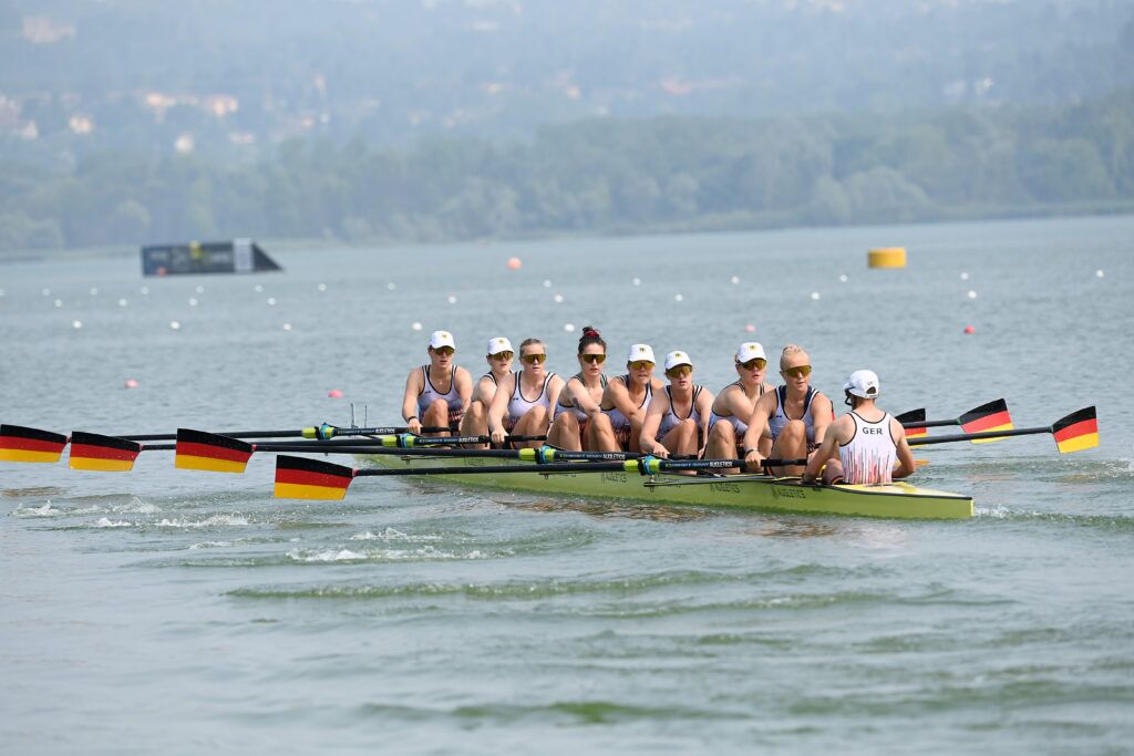 Der deutsche Frauen-Achter beim Rudern auf einem Regattakurs mit Deutschlandflaggen an den Ruderblättern und AUGLETICS-Branding auf dem Boot.