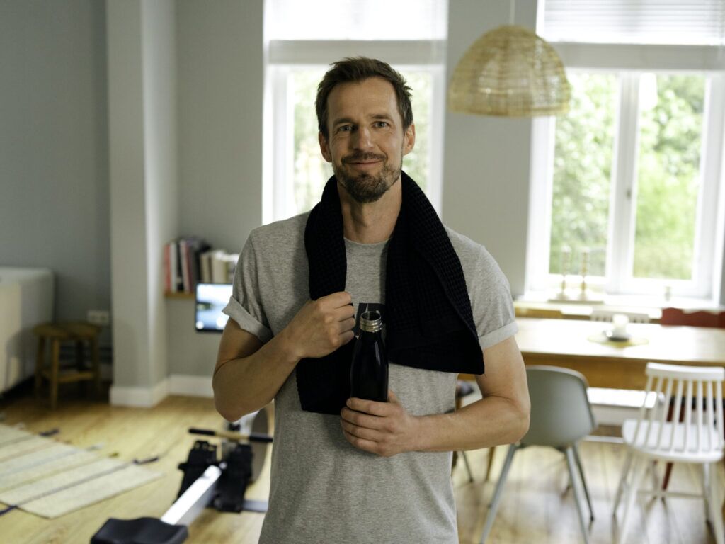 Mann mit Handtuch und Wasserflasche nach dem Training in einem Wohnzimmer mit Rudergerät.